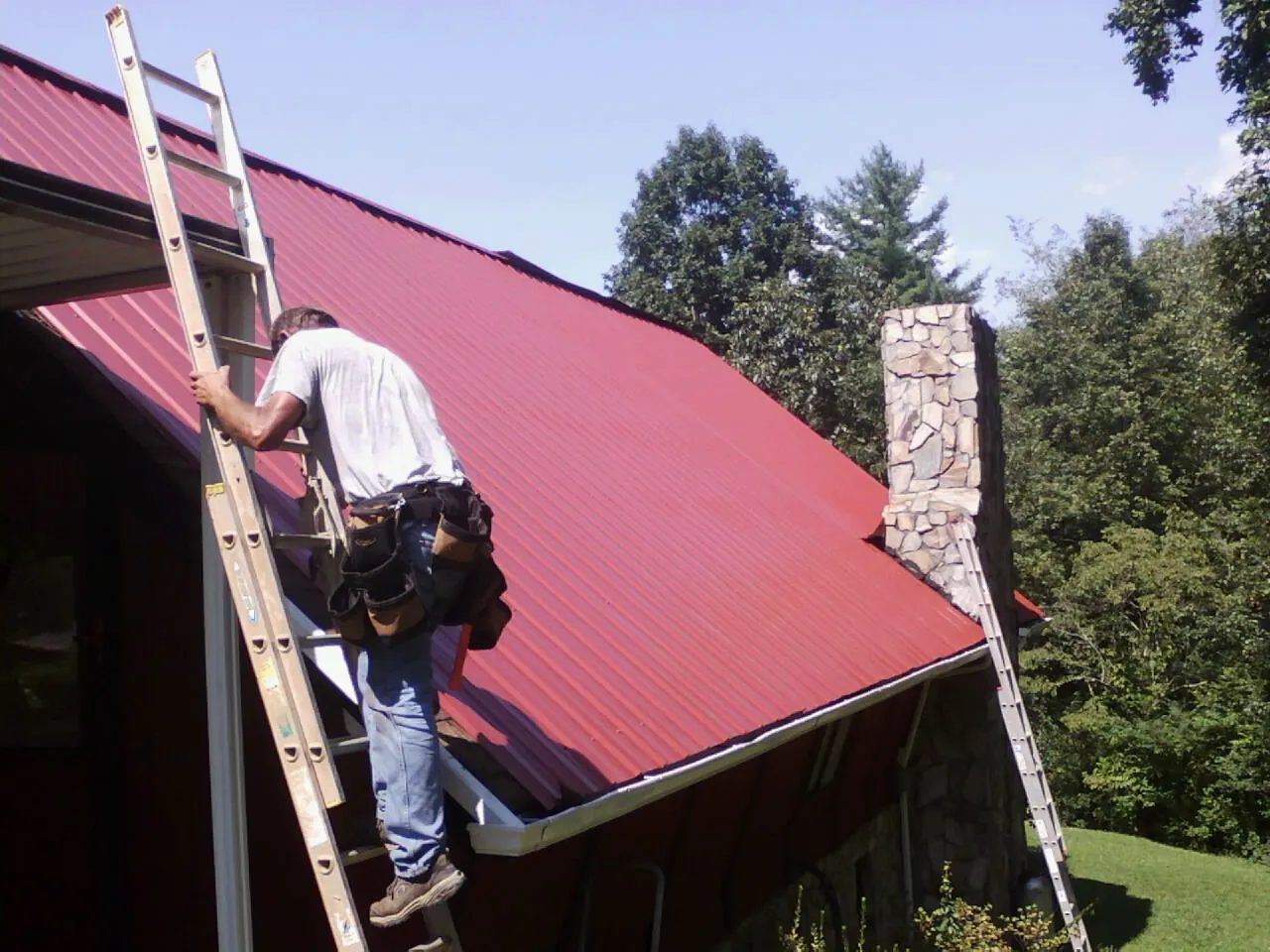 Roofer on a ladder beside a red metal roof and stone chimney — Soffit & Fascia Repair by Roofing Brothers in Winterthur